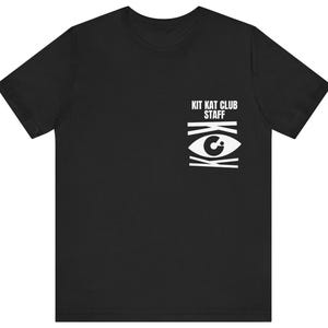 May include: Black t-shirt with the text "KIT KAT CLUB STAFF" above a white graphic eye design. The eye design is composed of horizontal and vertical lines, creating a unique visual element. The shirt is a classic crew neck style.