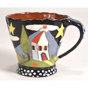 Könnte beinhalten: Handbemalte Keramiktasse mit einem skurrilen Landschaftsdesign. Die Tasse hat eine schwarze Außenseite mit einem weißen Haus, einem Baum und gelben Sternen. Der Boden hat ein weißes Tupfenmuster, und der Innenraum ist braun.