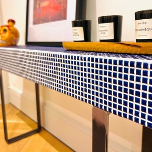 Puede incluir: Una mesa consola de azulejos azules y blancos con patas de metal negro. La mesa tiene una tapa de madera con tres velas encima. Las velas están etiquetadas como "Burning Rose", "Morning Rose" y "Bibliotheque".