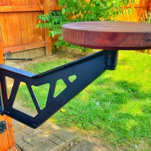 May include: A black metal bracket with a triangular design is attached to a wooden fence. A round wooden table top is mounted to the bracket.