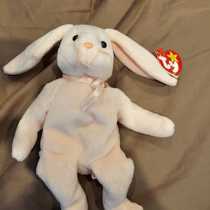 May include: A pink plush bunny with a white belly and long ears. The bunny is wearing a pink ribbon around its neck. The bunny has a red Ty tag attached to its ear that says "Ty Inc."