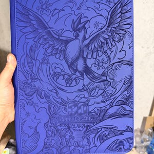Op de afbeelding: Een rechthoekige paarse map met een donkerblauwe reliëfafbeelding van een vogel met gespreide vleugels, omringd door wervelende wolken en sneeuwvlokpatronen. De tekst 30 ANNIVERSARIO staat onderaan met een klein silhouet van een Pikachu.