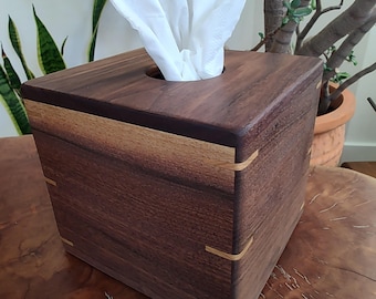 May include: A square wooden tissue box with a dark brown finish and light wood accents. White tissues are pulled from the circular opening on top. The box is sitting on a wooden surface.