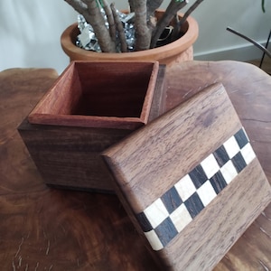 May include: A wooden box with a lid, crafted from dark brown wood. The lid features a decorative checkerboard pattern in black and white. The interior of the box is a lighter shade of brown. The box is open, revealing the inside.
