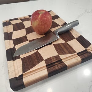 May include: A wooden chopping board with a chequered pattern, a groove around the edge, a stainless steel chef's knife, and a red apple. The board's pattern is made of light and dark wood squares.