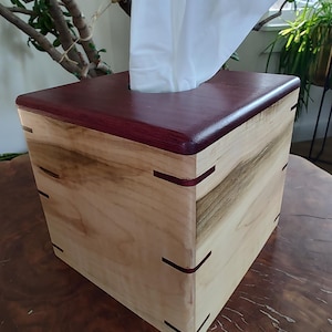 May include: A wooden tissue box with a light-colored wood body and a dark burgundy top. The box has a square shape with visible joinery and a contrasting dark wood accent. White tissues are visible.