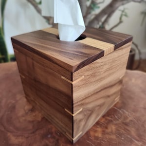 May include: A wooden tissue box with a square shape. The box is made of dark brown wood with lighter wood accents on the top and corners. White tissues are visible, emerging from a circular opening on top. The box is sitting on a wooden surface.