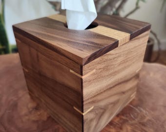 May include: A wooden tissue box with a square shape. The box is made of dark brown wood with lighter wood accents on the top and corners. White tissues are visible, emerging from a circular opening on top. The box is sitting on a wooden surface.