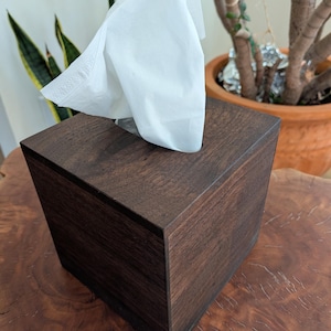 May include: A square tissue box cover made of dark brown wood with a visible grain pattern. A single white paper tissue is pulled through the rectangular opening at the top, extending upwards from the center of the wooden container.