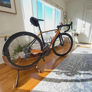 以下が含まれることがあります： オレンジと黒のSpecializedロードバイクが木製の自転車スタンドに展示されています。自転車は黒いホイール、黒いシート、黒いハンドルバーを備えています。自転車スタンドは木製で、自転車のホイールを保持する湾曲したデザインになっています。