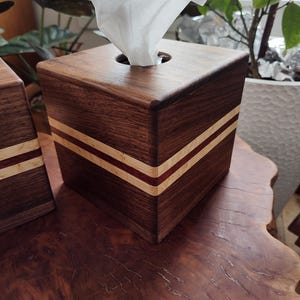 May include: A wooden tissue box cover with a dark brown finish and two thin red and white stripes around the top edge. The box is sitting on a wooden surface.
