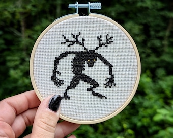 May include: A black and white cross-stitch embroidery hoop with a design of a creature with antlers and glowing eyes. The creature is stitched in black thread on a white background.