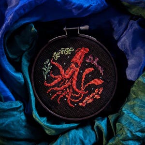 May include: A black embroidery hoop with a red octopus design on a black background. The octopus is surrounded by small, colorful flowers and stars.