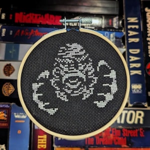 May include: A black and white cross-stitch embroidery of a creature from the movie 'The Creature from the Black Lagoon' in a wooden embroidery hoop.