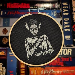 May include: A black and white cross-stitch embroidery hoop featuring a stylized image of a creature with large ears and a pointed head. The creature is wearing a white shirt and has a dark background.