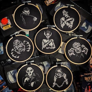 Könnte beinhalten: Sechs schwarze Stickrahmen mit weißen Stickereien von klassischen Horrorfilmfiguren. Die Figuren sind Frankensteins Monster, Dracula, die Kreatur aus der Schwarzen Lagune, der Werwolf und zwei weitere.