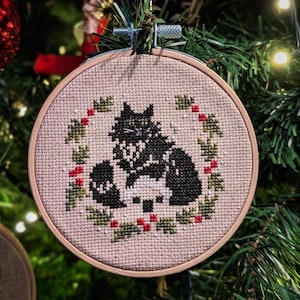 Peut inclure: Une décoration de Noël en tissu beige brodée d'un chat noir avec des marques blanches. Le chat est entouré d'une couronne de feuilles de houx vertes et de baies rouges. La décoration est circulaire et possède un cadre en bois.