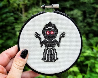 May include: A white embroidery hoop with a black and white cross-stitch design of a creature with red eyes. The creature has arms outstretched and is wearing a long black dress.