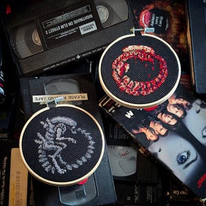 May include: Two cross-stitch hoops with horror movie designs. One has a red and black pattern, the other a grey alien on black. They are placed on VHS tapes, including "Nightmare on Elm Street 3."