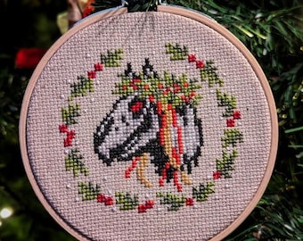 May include: A white and gray dinosaur with red eyes wearing a green holly wreath with red berries. The dinosaur is embroidered on a beige fabric with a wooden hoop.