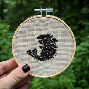 以下が含まれることがあります： ベージュの布地に、スタイリッシュな魚が黒と白のクロスステッチで刺繍されています。布地は木製の輪っかで張られています。