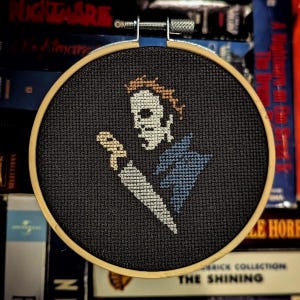 May include: A black and white cross-stitch embroidery of a horror movie character holding a knife. The character is wearing a blue and white shirt and has a white mask. The embroidery is framed in a wooden hoop.