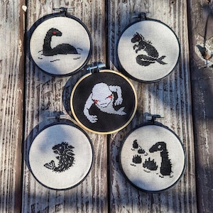 May include: Five embroidery hoops with black and white embroidered images of mythical creatures. The images include a Nessie-like creature, a ghost, a creature with a long tail, a creature with a long neck, and a creature with a long tail and a crown.