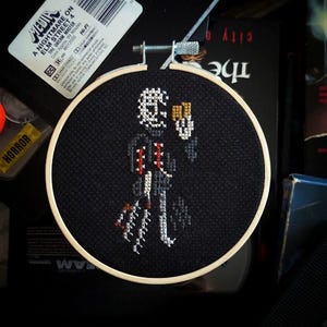 May include: A black and white cross-stitch embroidery of Freddy Krueger from the movie 'A Nightmare on Elm Street'. The embroidery is framed in a wooden hoop and shows Freddy holding a knife.