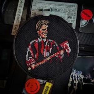 Könnte beinhalten: Eine schwarze und rote Kreuzstich-Stickerei eines Mannes, der eine Axt hält, mit Blut, das von der Axt tropft. Der Mann trägt ein weiß-rot gestreiftes Hemd. Die Stickerei ist in einem runden Holzrahmen gerahmt.