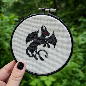 May include: Black and white cross-stitch embroidery of a small, winged creature with red eyes. The embroidery is framed in a round, black hoop.