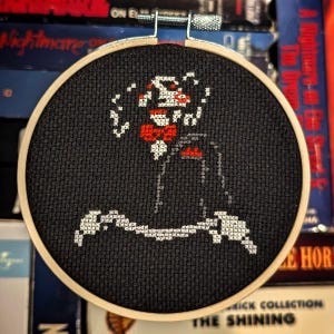 May include: A black and white cross-stitch embroidery of a character from the horror movie "The Shining" in a round wooden hoop. The character is wearing a black suit and a red bow tie. The background is black.