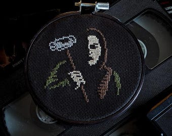 May include: A black and white embroidery hoop with a cross-stitch design of a hooded figure holding a flower. The figure is wearing a brown and white patterned robe. The background is black.