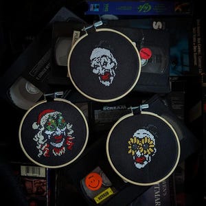 May include: Three embroidered hoop art pieces featuring different creepy clown faces. The hoops are black with a natural wood frame. The clowns are all white with red lips and black eyes. One clown is wearing a Santa hat and has green eyes. Another clown has yellow eyes and is wearing a yellow and black striped hat. The third clown has a skull-like face and is wearing a black hat.