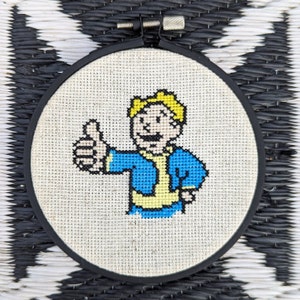May include: A cross-stitch embroidery of a cartoon character with blonde hair, wearing a blue jacket and yellow shirt, giving a thumbs up. The embroidery is framed in a black circular hoop.
