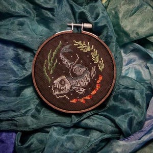 May include: A dark brown wooden embroidery hoop with a black and white cross-stitch design of a fish swimming in a dark blue sea. The hoop is surrounded by a teal blue fabric.