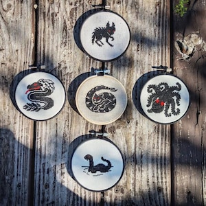 May include: Five embroidery hoops with black and white cross-stitch designs of mythical creatures. The designs include a snake, a monster, an octopus, a sea serpent, and a creature with large eyes.