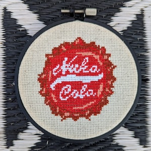 May include: A cross-stitch embroidery in a round black hoop. The design features a red and white "Kuka Cola" logo, reminiscent of a soda bottle cap, stitched on a beige fabric. The background is a black and white woven pattern.