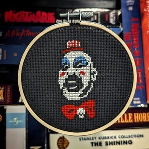 Puede incluir: Un bastidor de bordado de punto de cruz con la cara de un payaso con una pajarita roja y una calavera. El payaso tiene la piel blanca, maquillaje azul en los ojos y un sombrero rojo y blanco. El fondo es negro.
