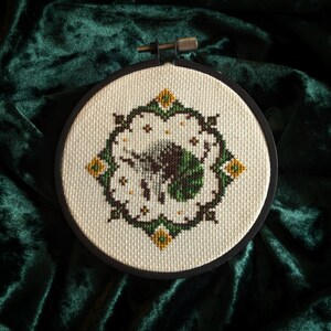 May include: A circular cross-stitch embroidery piece with a floral pattern. The design uses white, green, brown, and yellow threads in a symmetrical arrangement. The embroidery is on cream fabric, held within a black embroidery hoop.