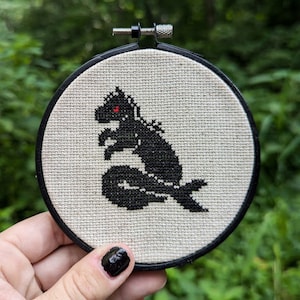 May include: A black and white cross-stitch embroidery of a Godzilla-like monster in a circular hoop.