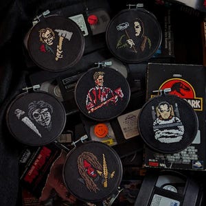 Könnte beinhalten: Sechs schwarze Stickrahmen mit bunten Kreuzstich-Designs von Horrorfilmfiguren. Die Figuren sind Jason Voorhees, Freddy Krueger, Michael Myers, Leatherface, Ghostface und Chucky.