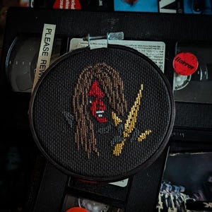 Op de afbeelding: Een zwarte borduurring met een kruissteekontwerp van een rood gezicht met lang bruin haar en een gouden mes. Het ontwerp is op een zwarte achtergrond. De ring ligt op een stapel VHS-banden.