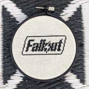 May include: A white embroidery hoop with black embroidery of the word "Fallout" in a rectangular frame.