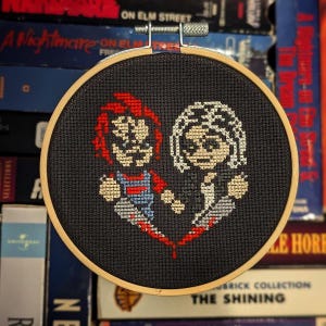 May include: Cross-stitch embroidery hoop with a black background and a red heart design. The heart features a pixelated image of Freddy Krueger and Jason Voorhees from the horror movies A Nightmare on Elm Street and Friday the 13th.