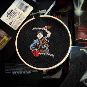 Puede incluir: Un aro de bordado de punto de cruz en blanco y negro con una imagen pixelada de Freddy Krueger de la película "A Nightmare on Elm Street". El aro está rodeado de cintas VHS.