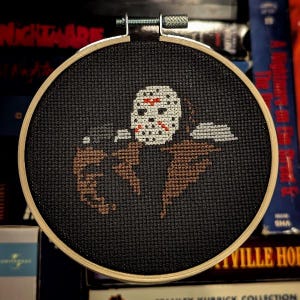 May include: A black and white cross-stitch embroidery of Jason Voorhees, the hockey mask-wearing killer from the Friday the 13th horror franchise. The embroidery is framed in a wooden hoop.