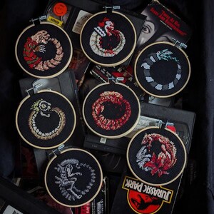 May include: Eight embroidered hoops with black fabric and colorful thread depicting iconic horror movie monsters. The hoops are arranged in a circle around a stack of VHS tapes. The movie titles "Scary and Entertaining", "Toner As Hie", "JURASSIC PARK", and "YOUR WILL" are visible on the tapes.