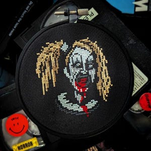 May include: A black embroidery hoop with a cross-stitch design of a zombie with long, blonde hair and a bloody mouth. The zombie is stitched in shades of white, gray, and red.
