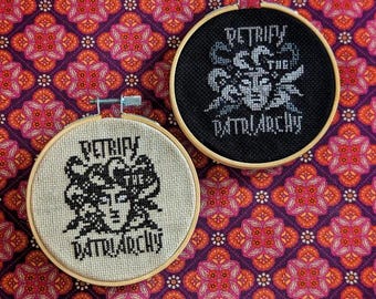 May include: Two embroidery hoops with black and white cross-stitch designs. The designs feature a woman's face with snakes and the text "Petrify the Patriarchy".