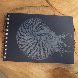 May include: A black spiral bound notebook with a silver nautilus shell illustration on the front cover.